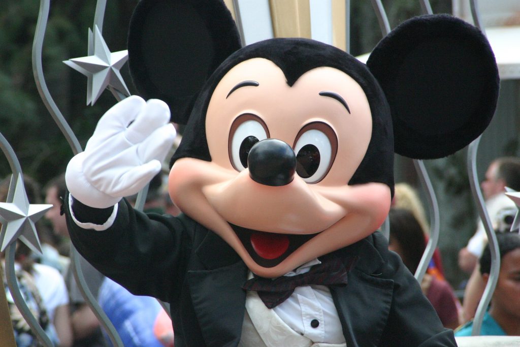 Mickey Mouse, dressed in a black dinner jacket with a bow tie, waves and smiles at the camera. Silver star decorations and people in the background suggest a festive California parade setting.