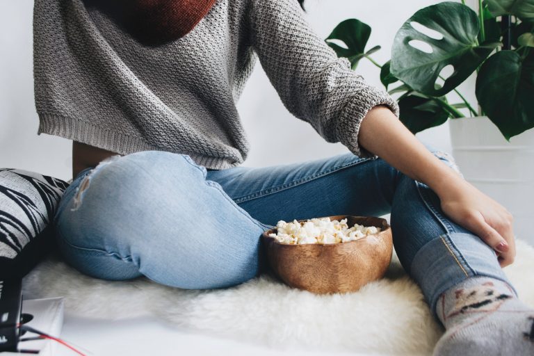 For a cosy girls' night in, a person in a grey jumper and blue jeans sits cross-legged on a fluffy white rug, reaching for popcorn in a wooden bowl. A green plant adds freshness to the scene, with only the lower half of the person visible.