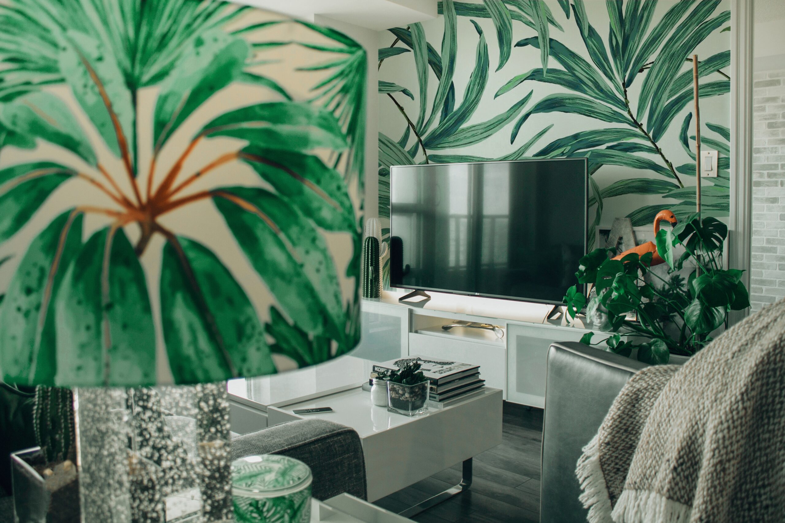 A modern lounge with leafy green wallpaper, a TV on a white stand, plants, and a lamp with a green leaf-patterned shade in the foreground showcases one of the popular interior design styles with lush, tropical vibes.