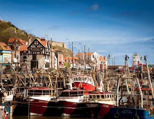 A busy harbour filled with colourful fishing boats, charming buildings, and shops along the waterfront—this vibrant scene is one of the Great Destinations in the UK for Weekend Breaks, complete with a Ferris wheel and cliffs under a bright blue sky.
