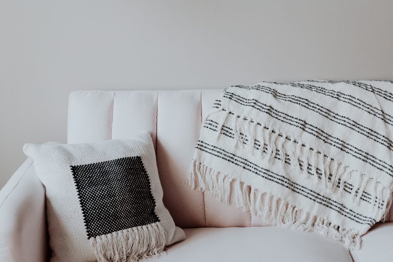a cushion on a cream sofa with a grey and white throw on the back