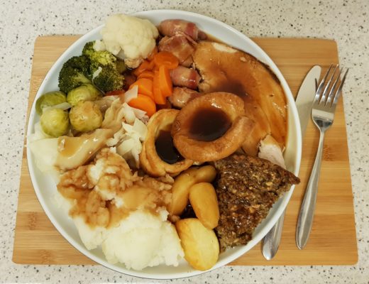 A plate of quick and easy Christmas dinner featuring sliced meat, gravy, roast and mashed potatoes, Yorkshire pudding, stuffing, carrots, cabbage, broccoli, cauliflower, Brussels sprouts, and pigs in blankets on a wooden board with cutlery.