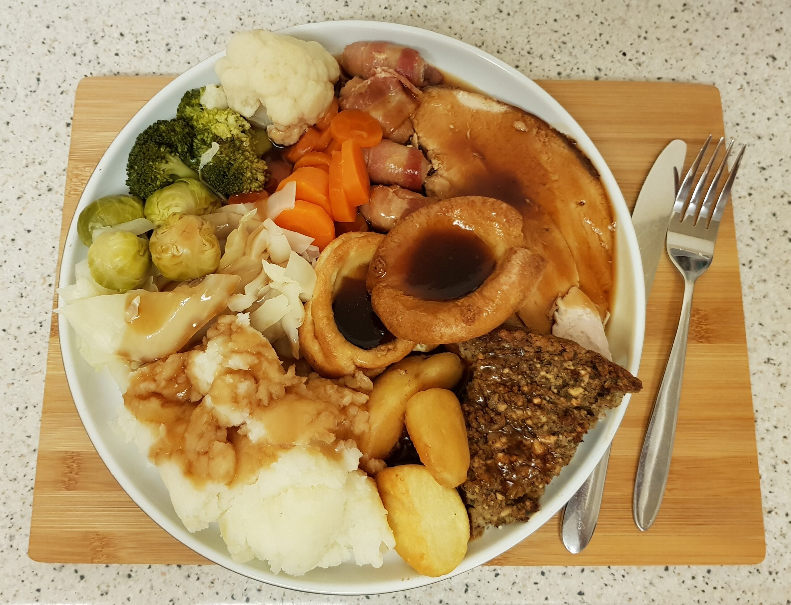 A plate of quick and easy Christmas dinner featuring sliced meat, gravy, roast and mashed potatoes, Yorkshire pudding, stuffing, carrots, cabbage, broccoli, cauliflower, Brussels sprouts, and pigs in blankets on a wooden board with cutlery.