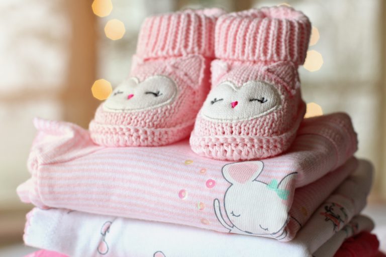 A pair of pink knitted baby booties with a sleeping owl design rests on a neatly folded stack of pink and white baby clothes, evoking cosy warmth—an ideal scene for sharing parenting tips with new mothers.