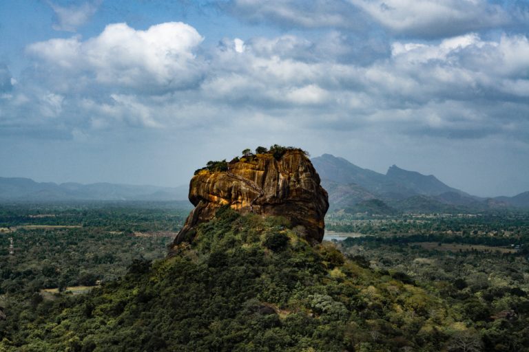 A large, rocky outcrop rises above dense green forest under a partly cloudy sky, with distant mountains visible in the background—the perfect setting for a secluded luxury holiday.