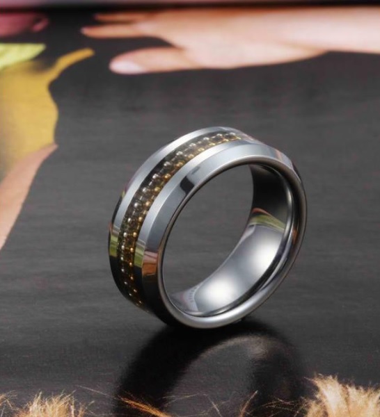A silver ring with a gold stripe, featuring small embedded stones, stands upright on a glossy, blurred magazine page. The elegant design echoes the modern style of Tungsten Carbide Rings amid subtle hints of skin tones and colours in the background.