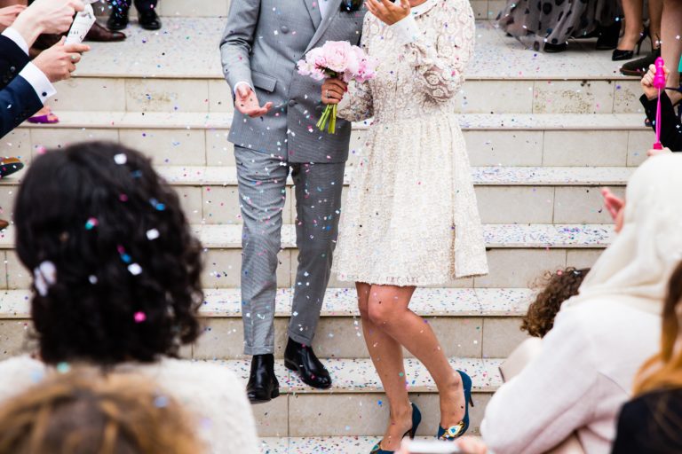 A couple, dressed in wedding attire, walk down outdoor steps at their picturesque waterfall wedding as guests throw confetti. The bride holds pink flowers and smiles, while the groom in a grey suit beams amid the festive, joyful scene.