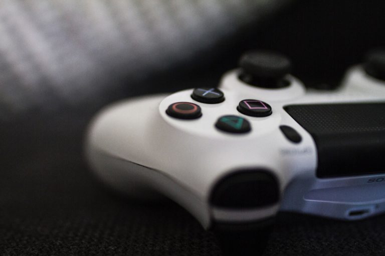 Close-up of a white PlayStation controller resting on a dark surface, focusing on the coloured shape buttons and thumbsticks.