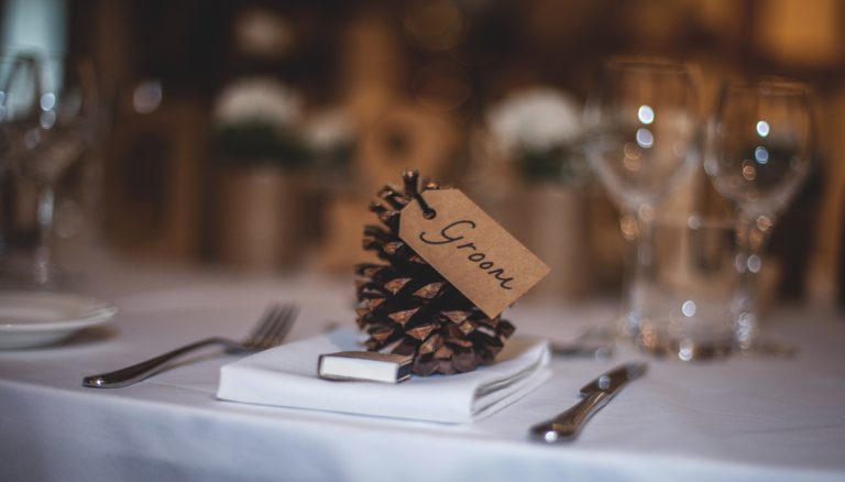 A pine cone with a "Groom" tag serves as a place card, resting on a folded white serviette at a formal table setting with cutlery, plates, and wine glasses.
