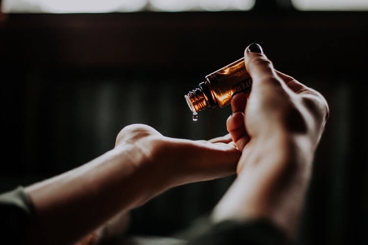 A person pours natural oil from a small amber glass bottle onto their palm, with a blurred dark background.