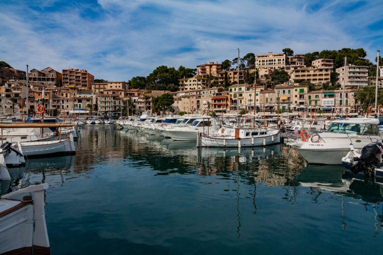 Boats moored in a marina with calm water, surrounded by colourful buildings and green hills under a partly cloudy sky. The scene is lively and picturesque, evoking a Mediterranean coastal town atmosphere.