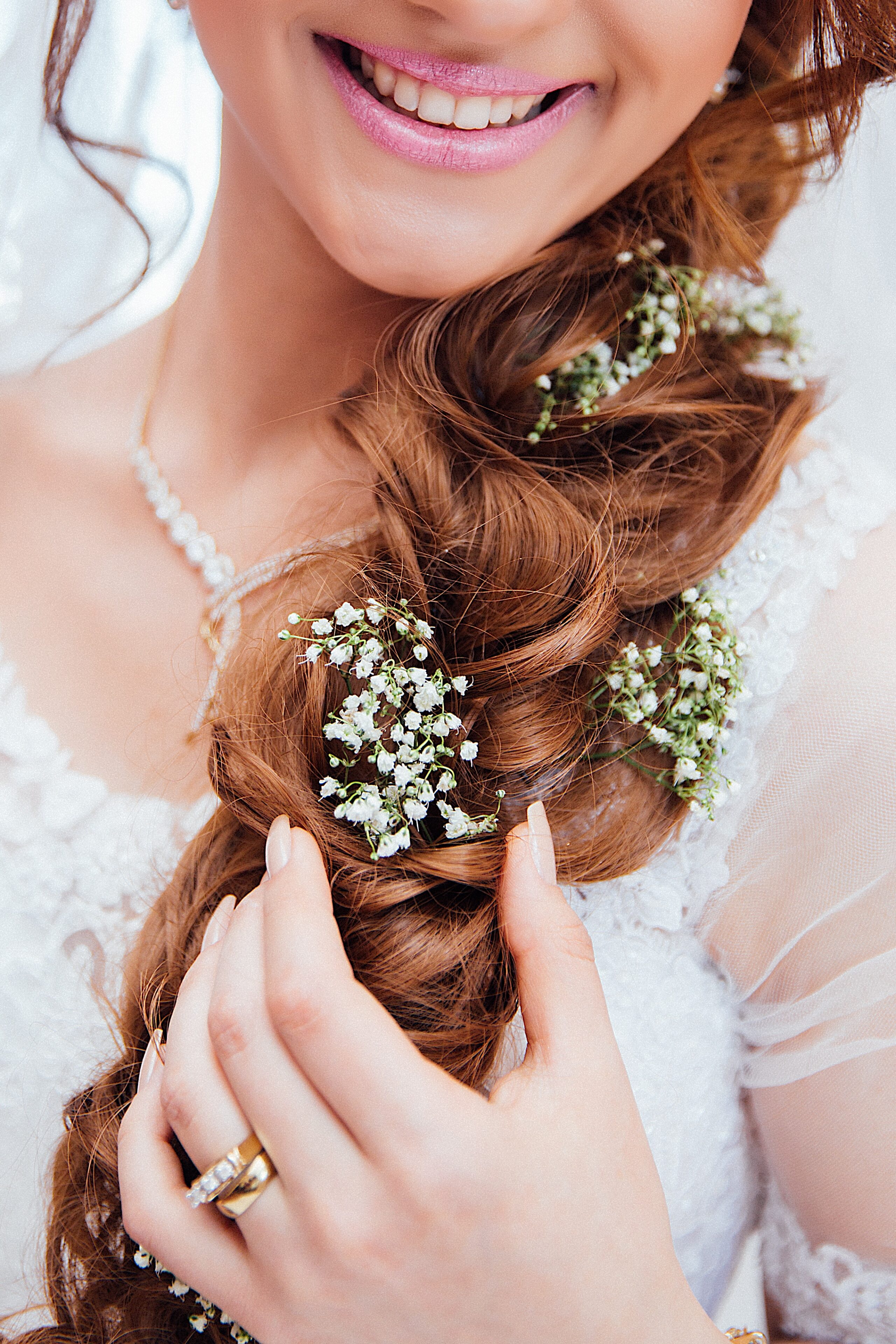 A close-up of a smiling bride showcases bridal beauty with long auburn hair styled in a plait adorned with small white flowers. She wears a white lace dress, a pearl necklace, and a gold ring on her hand.