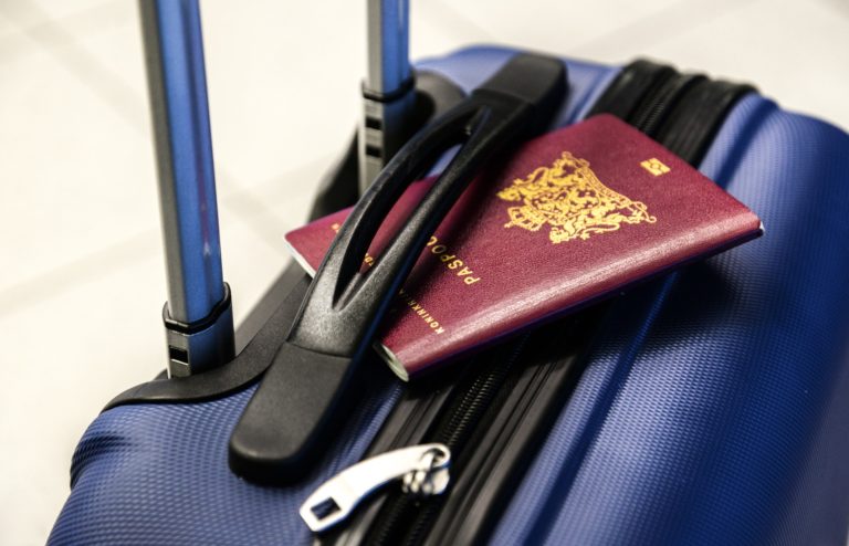 A maroon passport rests on top of a blue hard-shell suitcase with an extended handle and a visible zip, suggesting preparation for travel.