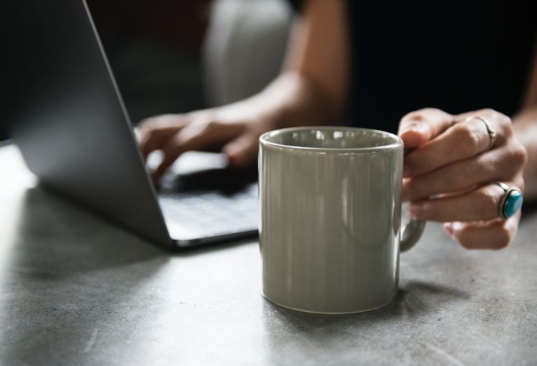 woman sat at a table workin on her laptop reaching for a white mug
