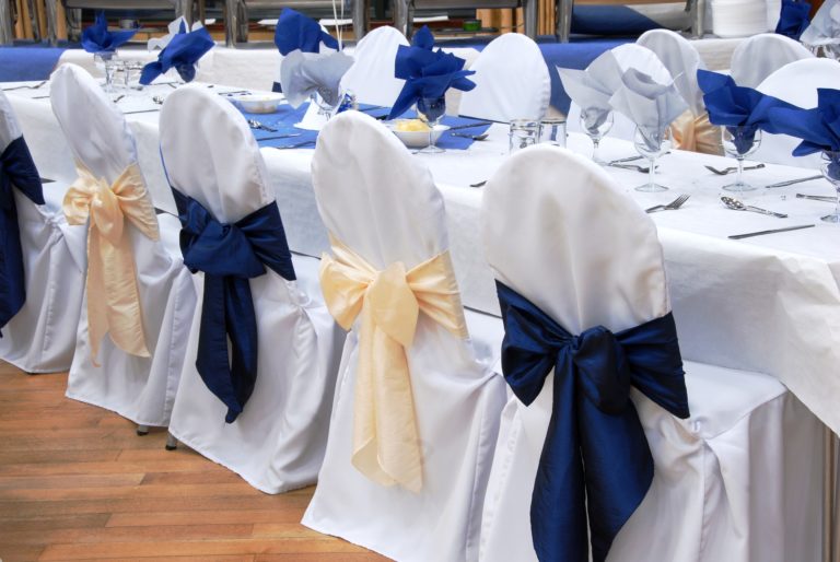 A long dining table set for an event with white tablecloth, white chairs with blue and cream bows, glasses, cutlery, and blue serviettes, on a wooden floor.