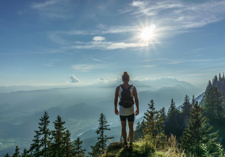 A person with a rucksack stands on a grassy mountain edge, gazing towards the future across vast hills and valleys. Pine trees frame the scene, and the sun shines brightly in a blue, partly cloudy sky.