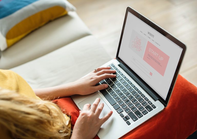 A person using a laptop, sitting on a sofa with colourful cushions. The screen displays a website titled "START UP" with a pink button, suggesting focus on entrepreneurship or launching an online business.