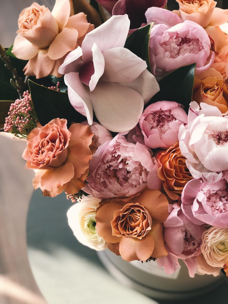 A must-have wedding preparation item, this bouquet features soft pastel flowers—pink peonies, peach roses, and white blooms—arranged closely together in a white vase for an elegant touch.
