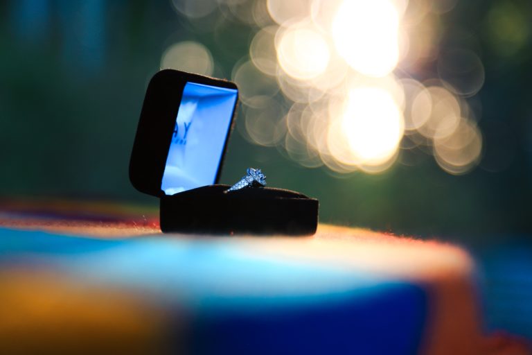 An engagement ring in an open black box is placed on a colourful surface, with warm, blurred lights and soft focus in the background.