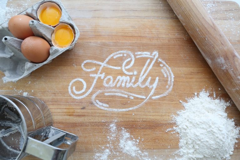 A wooden board with flour spelling out "Family", surrounded by eggs in a box, a pile of flour, a rolling pin, and a sieve—capturing the joy of baking as one of the best family hobbies.