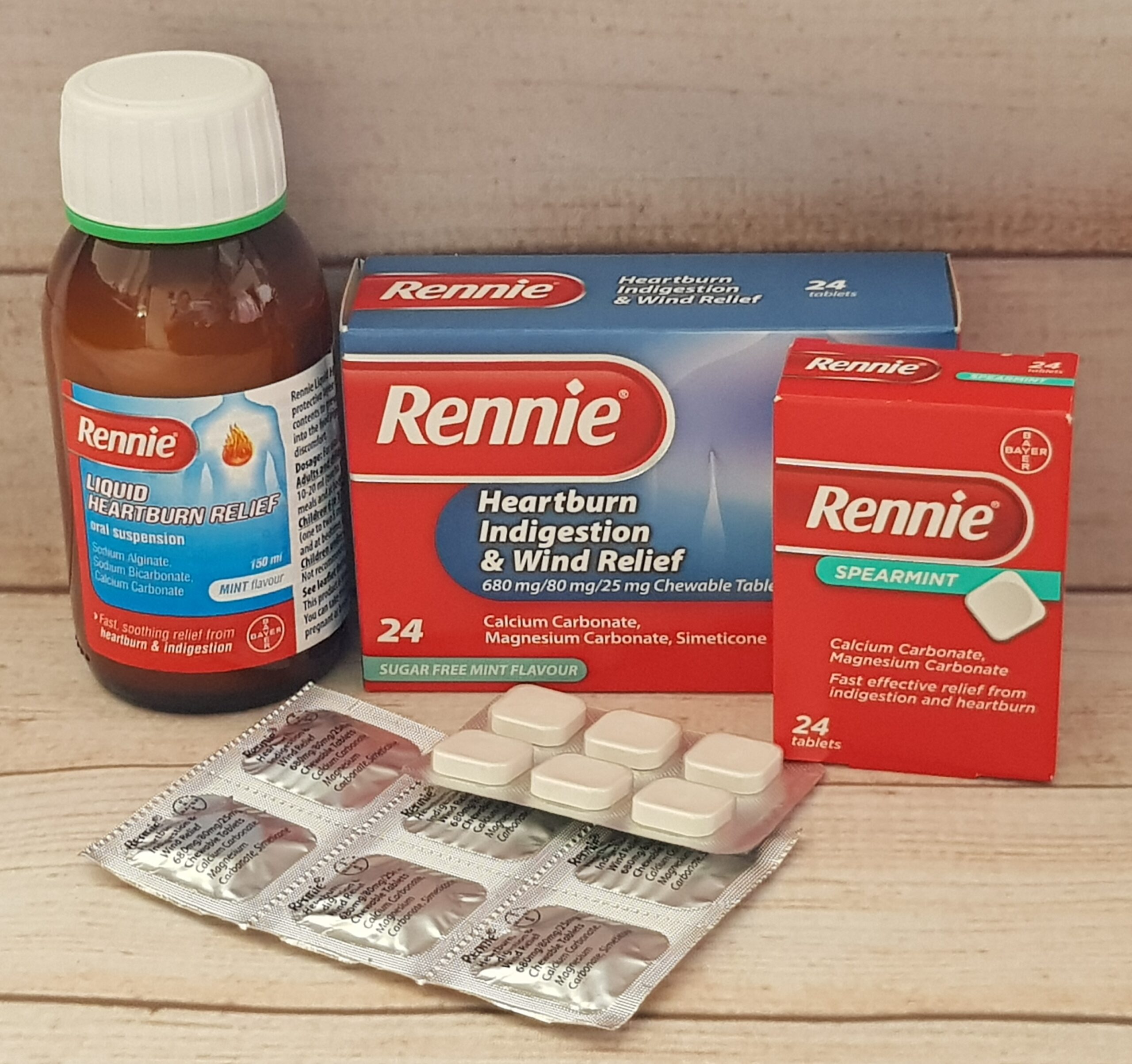 A bottle, two boxes, and blister packs of Rennie heartburn and indigestion relief medicine are displayed on a wooden surface. The boxes are labelled in red, white, and blue, with chewable tablets and liquid options shown.
