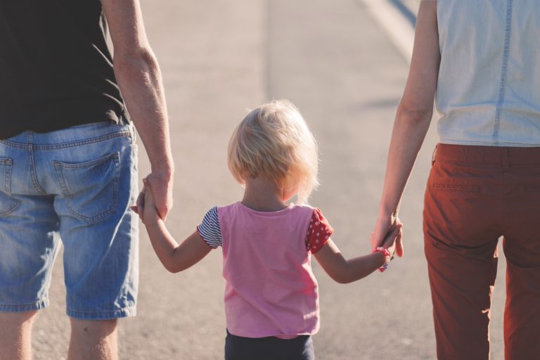 A young child with short blond hair holds hands with two adults, one on each side, as they walk together outdoors on a sunny family summer holiday. The photo is taken from behind.