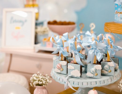 Decorative treats with blue bows and hot air balloon designs are displayed on a cake stand at a pastel-themed party table, surrounded by flowers and soft blue and white decorations.