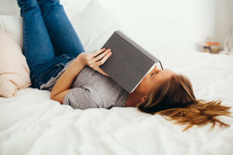 A woman lies on her back on a bed with white sheets, holding a closed book over her face. Wearing blue jeans and a grey t-shirt, she dreams of her upcoming wedding, legs bent at the knees and feet resting against the wall.