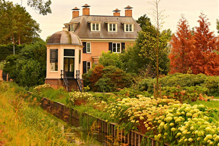 A large, elegant house perfect for selling a luxury home sits surrounded by lush green bushes, trees, and blooming flowers beside a small canal with a wooden fence along its edge.
