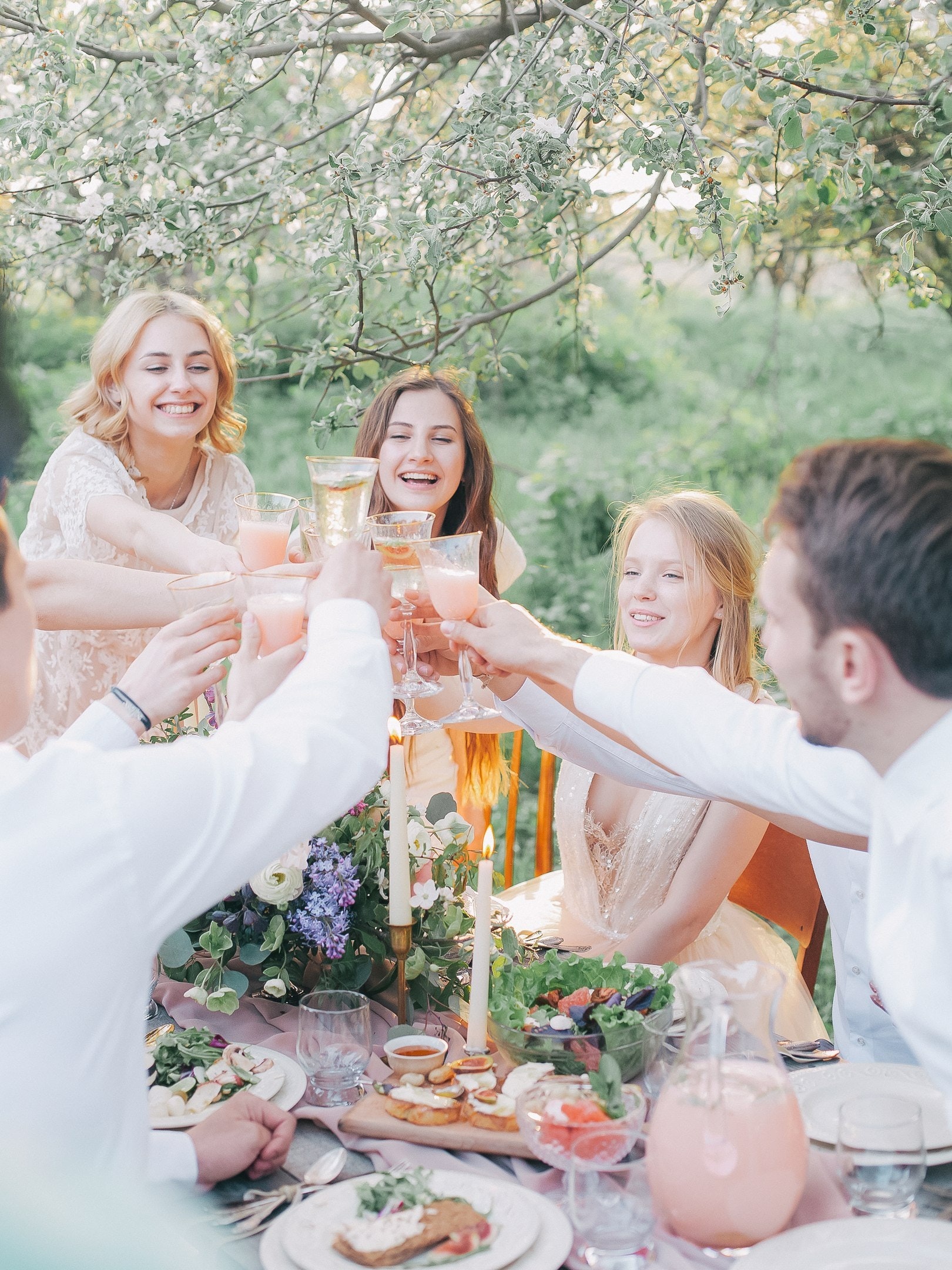A group of young adults, dressed in light-coloured clothes, sit outdoors around a decorated table, smiling and raising glasses for a toast at a summer party. The table is set with food, flowers, and candles under blossoming trees.