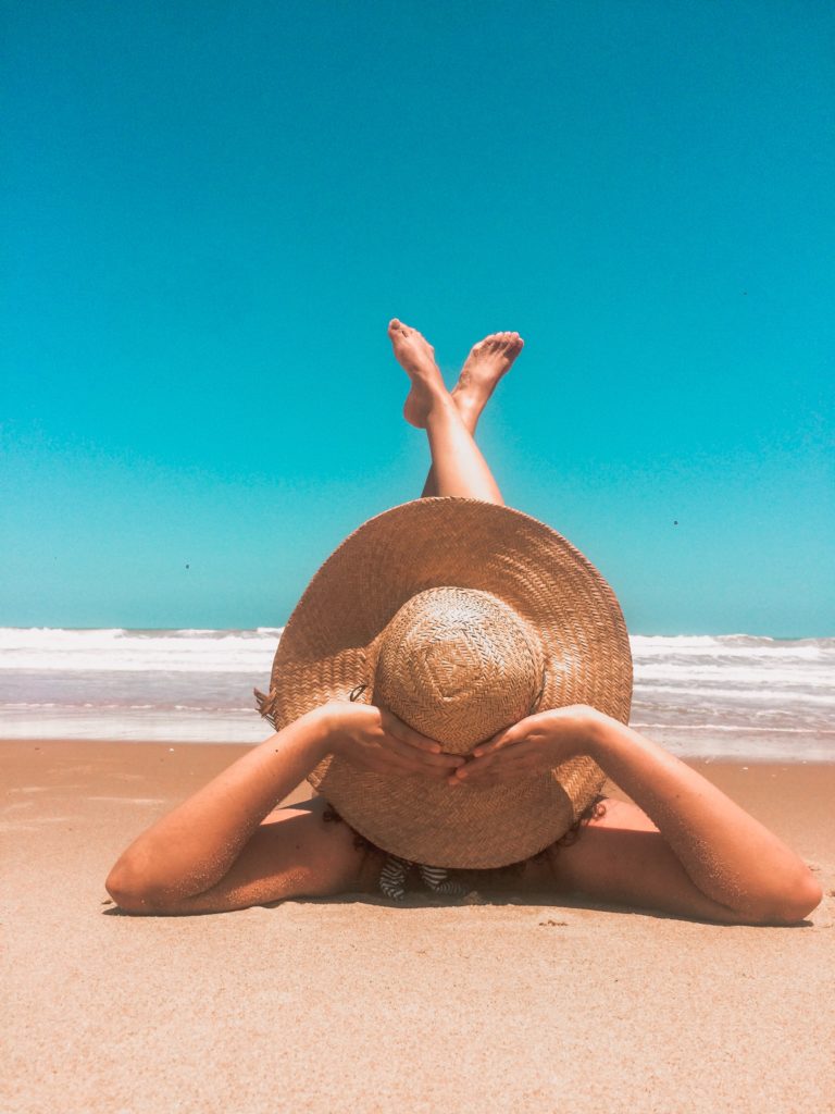 A person in stylish swimwear lies on a sandy beach with legs crossed in the air, wearing a wide-brimmed straw hat and facing the sea under a clear blue sky. The scene conveys relaxation and a sunny day at the seaside.