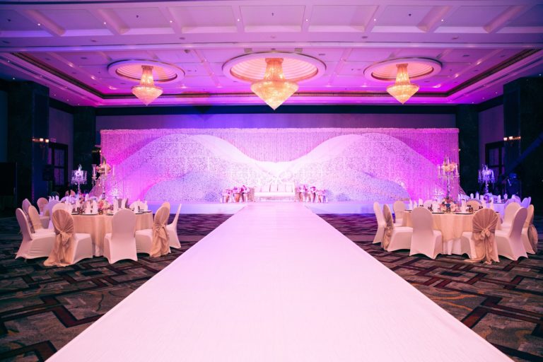A lavishly decorated banqueting hall, perfect for a post-elopement party, with round tables draped in white linen, elegant chairs, and a long white aisle leading to a stage with a floral backdrop and pink-purple lighting. Chandeliers hang from above.