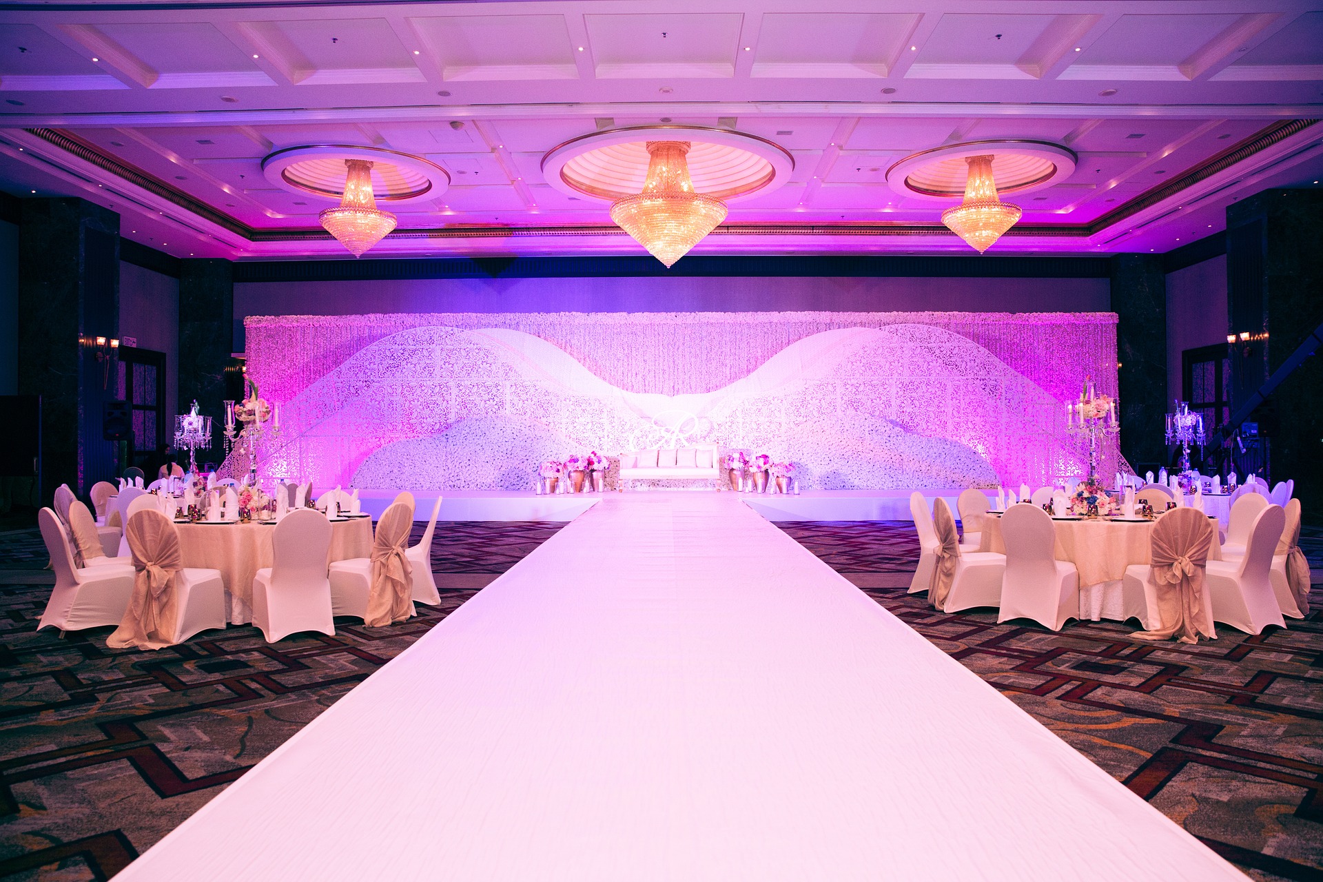 A lavishly decorated banqueting hall, perfect for a post-elopement party, with round tables draped in white linen, elegant chairs, and a long white aisle leading to a stage with a floral backdrop and pink-purple lighting. Chandeliers hang from above.