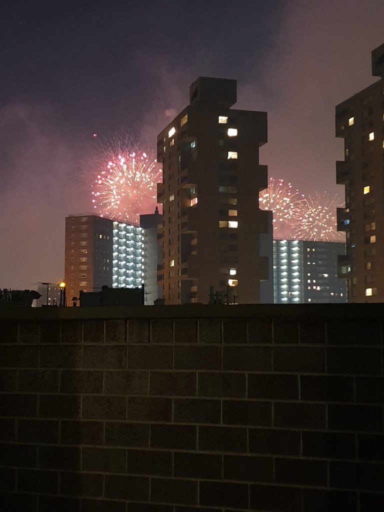 Fireworks light up the New York night sky behind tall blocks of flats, with window lights shining. A brick wall is in the foreground, and smoke from the fireworks drifts through the air.