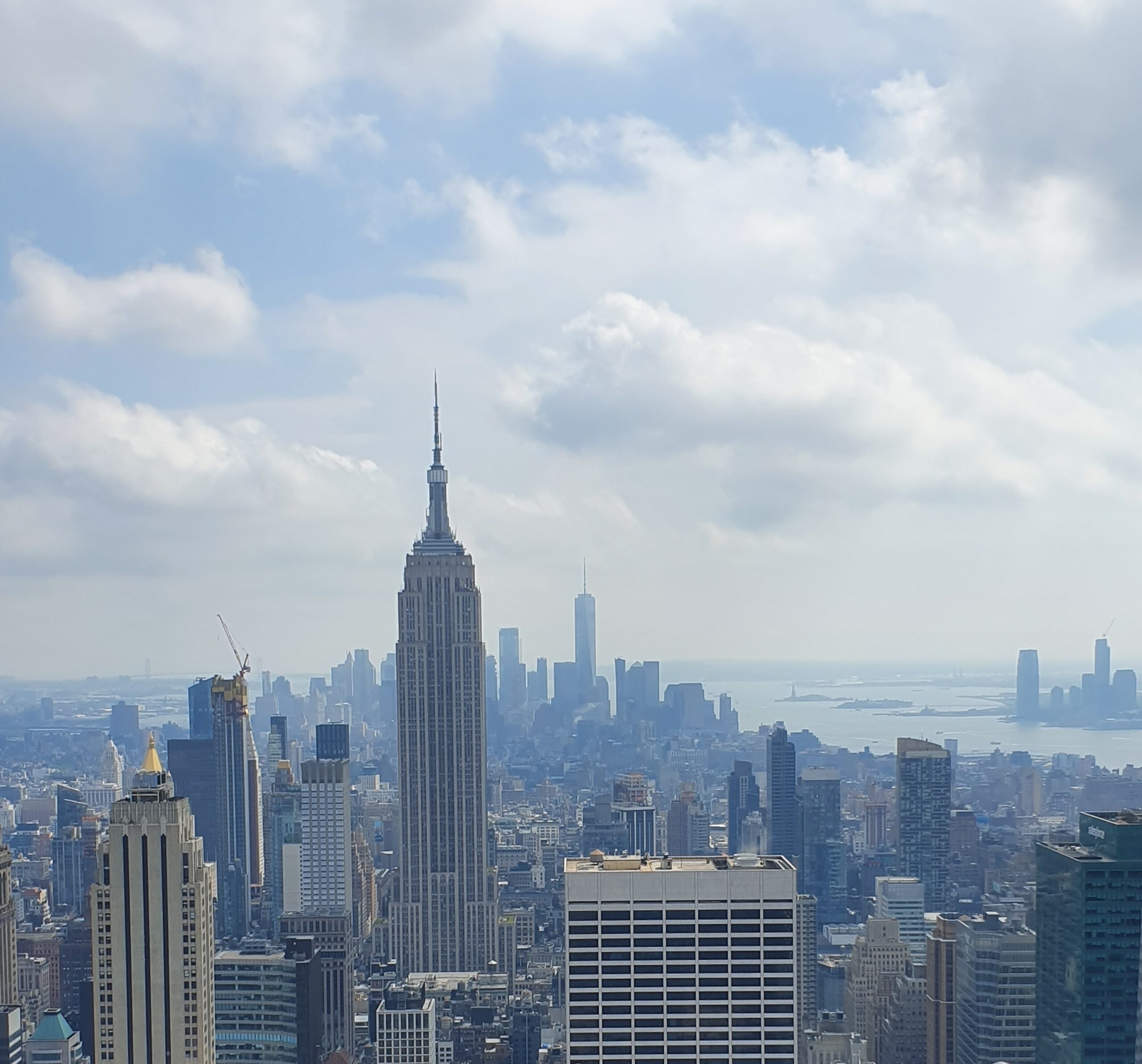 View of the New York City skyline featuring the Empire State Building in the centre, with other skyscrapers and cloudy sky above. The distant skyline and water are visible in the background.