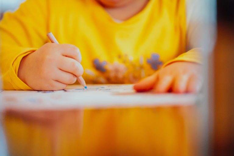 A prep school child wearing a yellow shirt is drawing or colouring on paper with a coloured pencil, focusing closely on their artwork. Their face is partly out of frame.