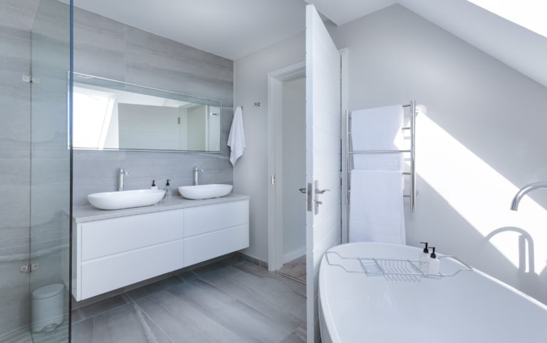 Modern bathroom with grey tiles, a double sink vanity with mirrors, a white freestanding bath, a towel rail with white towels, and natural light streaming in through a decorative glass window.