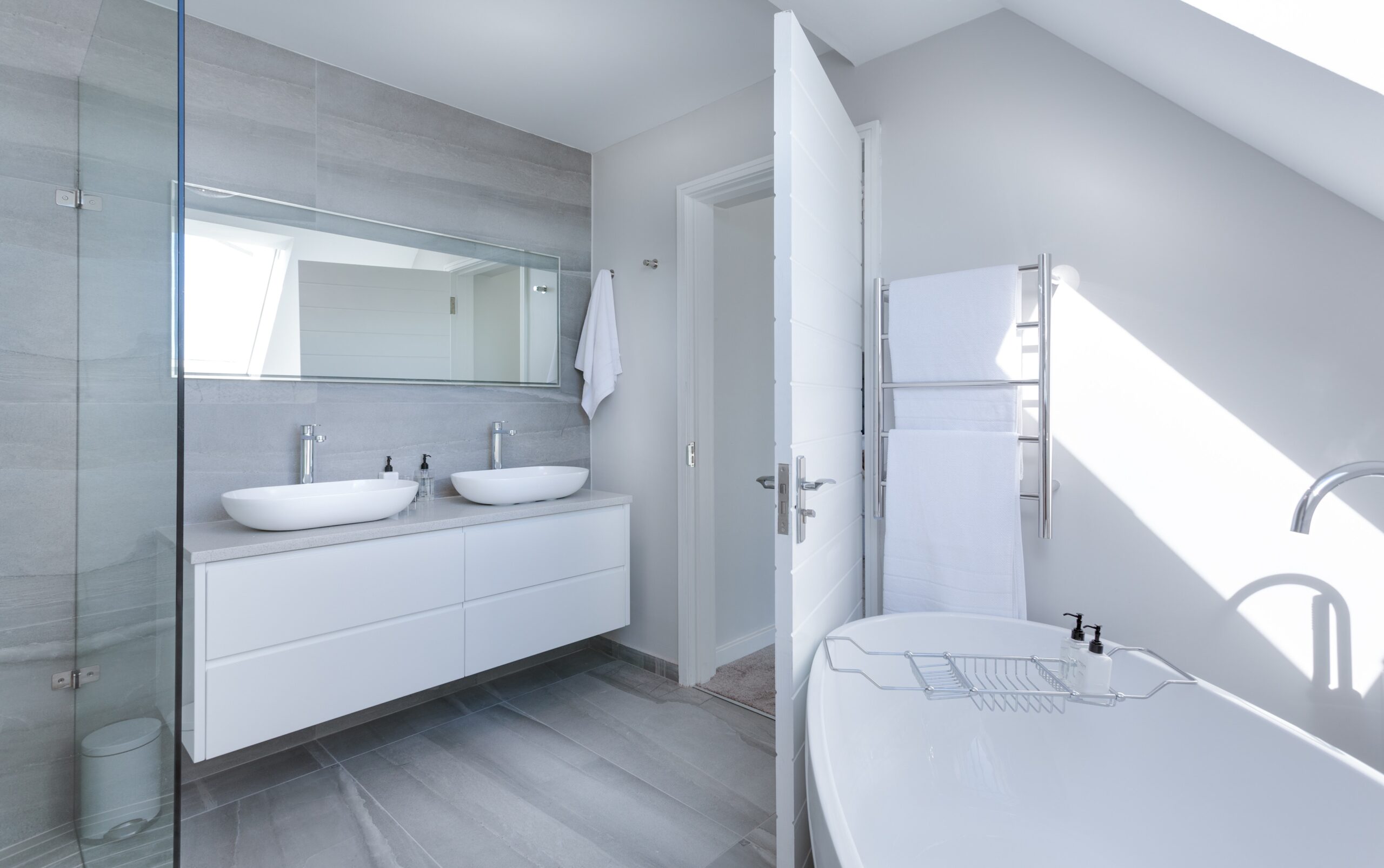Modern bathroom with grey tiles, a double sink vanity with mirrors, a white freestanding bath, a towel rail with white towels, and natural light streaming in through a decorative glass window.