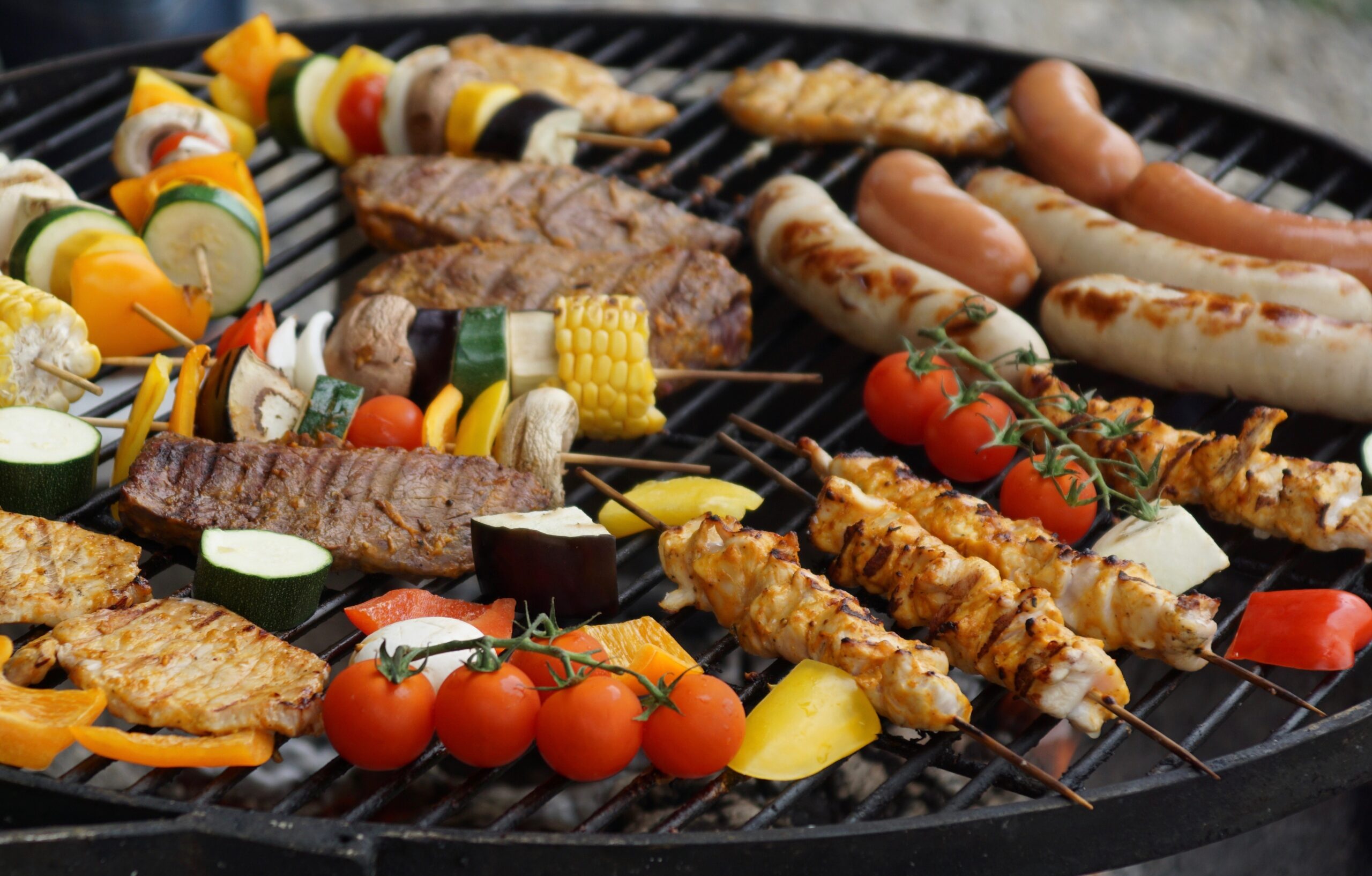 A variety of meats, sausages, chicken skewers, grilled vegetables, and cherry tomatoes sizzle on a round BBQ grill outdoors.