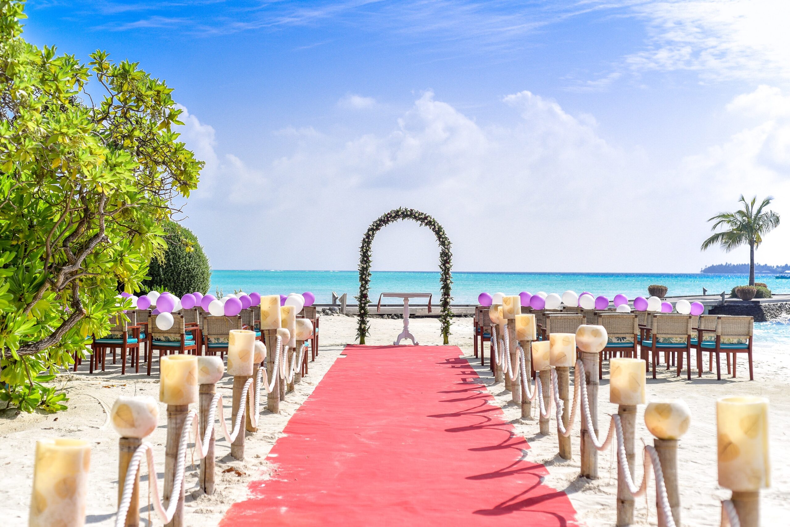 A memorable wedding set-up on the beach features a red carpet aisle lined with candles, leading to a floral arch. Chairs adorned with purple and white balloons face the sea beneath a bright blue sky dotted with clouds.