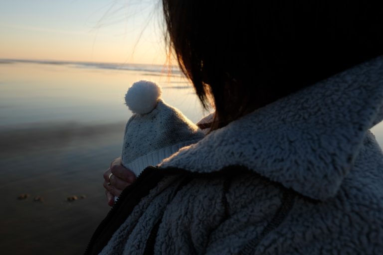 A person in a fluffy jacket holds a baby wearing a nursery-style knitted hat with a bobble, standing by the water at sunset. The scene is calm, with soft light reflecting on the water.