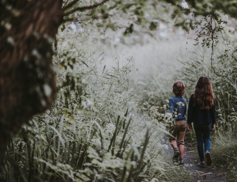 Two children, a boy and a girl, walk side by side on a narrow path through a lush forest, surrounded by tall grass and trees. Their calm child behaviour adds to the peaceful, natural scene.
