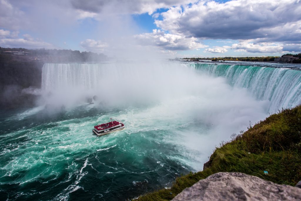 A tour boat approaches the base of Niagara Falls in Canada, surrounded by mist and powerful cascading water, with a rocky shore and green grass in the foreground under a partly cloudy sky.