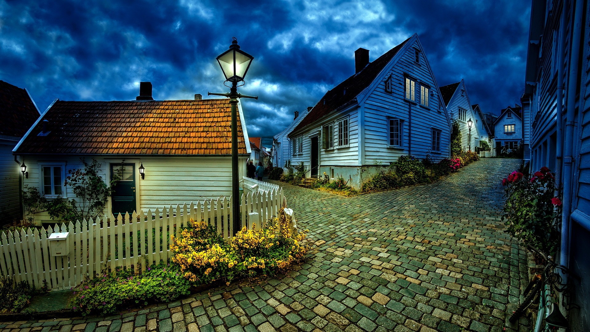 A cobblestone street lined with white wooden houses and a glowing streetlamp under a dramatic, cloudy evening sky evokes a sense of home. Yellow flowers and a white picket fence accent the cosy, inviting scene.