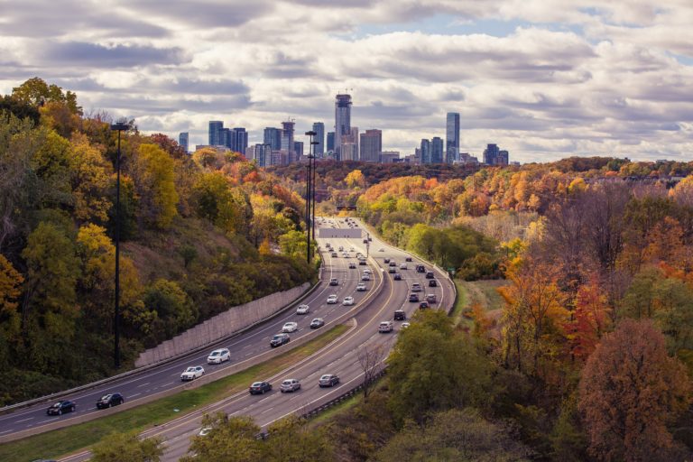 A city skyline in the distance under a cloudy sky, with a busy multi-lane motorway curving through a forested area of colourful autumn foliage, captures the vibrant beauty of Canada. Cars are travelling in both directions.