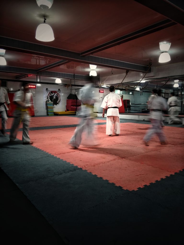 Several people in martial arts uniforms practise in a dojo with red mats and bright overhead lights. One person stands still at the centre—a moment that echoes the focus needed in the Five Things To Think About When Starting Up A Kid's Club.