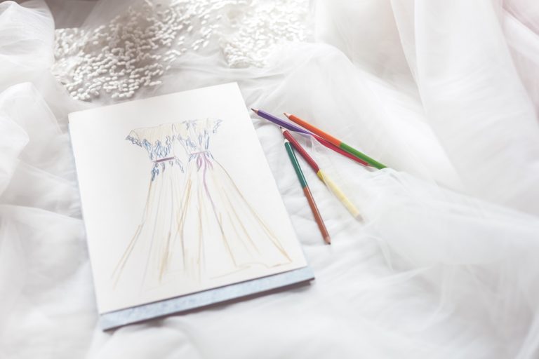 A sketch of a long, elegant dress on white paper lies on sheer white fabric, with five coloured pens—purple, orange, green, red, and yellow—placed beside it. White decorative beads and notes for wedding dress alterations are visible in the background.