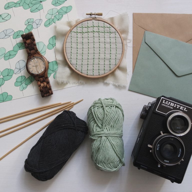 Flat lay of hobby craft supplies, featuring green and black wool, wooden knitting needles, an embroidery hoop with green stitching, a wooden watch, a vintage camera, envelopes, and leaf-patterned paper.