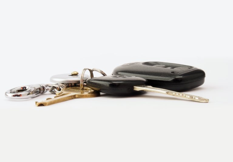 A set of keys on a keyring—including a car key with a black fob—suggests someone is buying a used car, alongside a brass house key and silver keyring, all arranged neatly on a white surface.