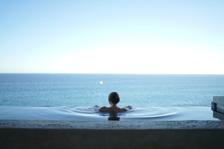 A person with hair in a bun relaxes in an infinity pool overlooking a calm, blue sea under a clear sky, perhaps planning their next trip or EHIC card renewal as the horizon meets the tranquil water.